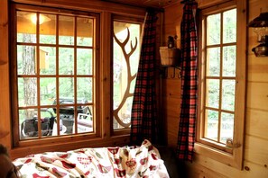 A cabin with large windows, a bed with a patterned blanket, plaid curtains, and a wooden interior. - Magical Tree House Rental in the Woods near Syracuse for Glamping in New York State (Lafayette)