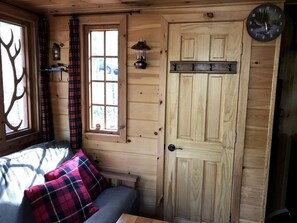 A wooden cabin interior with a bed, a window with curtains, a door, and a wall-mounted clock. - Magical Tree House Rental in the Woods near Syracuse for Glamping in New York State (Lafayette)