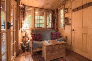 A cozy cabin interior with a sofa, a table with books, plaid curtains, and a wooden door. - Magical Tree House Rental in the Woods near Syracuse for Glamping in New York State (Lafayette)