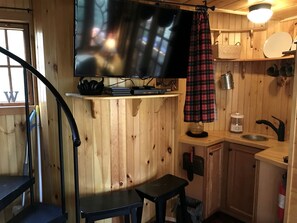 A wooden interior with a television, a small table, a bench, a kitchen area with a sink, and a staircase. - Magical Tree House Rental in the Woods near Syracuse for Glamping in New York State (Lafayette)
