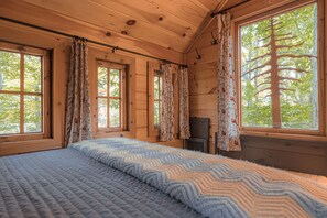 A wooden cabin room with a bed, large windows, and a fireplace. - Magical Tree House Rental in the Woods near Syracuse for Glamping in New York State (Lafayette)