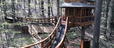 A wooden treehouse with a slide, surrounded by a forest.