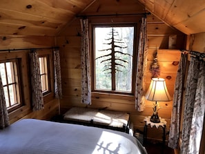 A wooden cabin room with a bed, a window with a tree view, a lamp, and curtains.