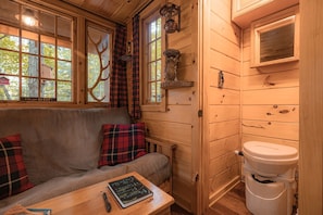 A wooden cabin interior with a sofa, plaid cushions, a table with a book, a window with a deer silhouette, and a toilet.