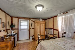 A bedroom with a wooden wardrobe, a bed with a patterned bedspread, a desk with a lamp, and a window with curtains.