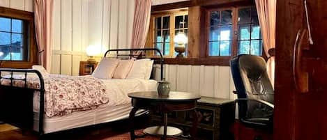 Una habitación acogedora con una cama, una mesa y una silla. Hay ventanas con cortinas, una lámpara y un techo de madera.