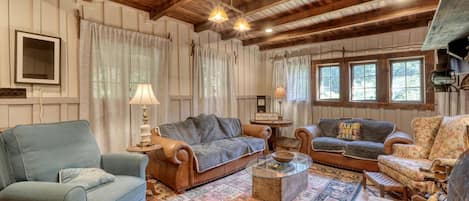 A cozy room with wooden beams, a patterned rug, and a variety of seating including a blue armchair, a brown sofa, and a patterned armchair.
