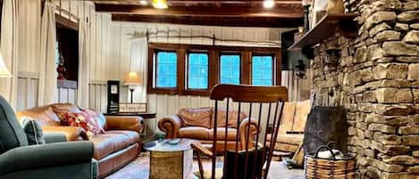Ein gemütliches Wohnzimmer mit Kamin aus Stein, Holzbalkendecke und einem großen Fenster mit vielen Scheiben.