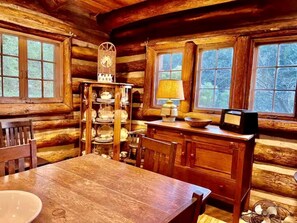 Ein rustikales Blockhäuschen im Inneren mit einem Holztisch, einer Uhr, einer Lampe und einem Regal mit Geschirr.