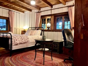 Une pièce confortable avec un lit, une table et une chaise. Il y a des fenêtres avec des rideaux, une lampe et un plafond en bois.