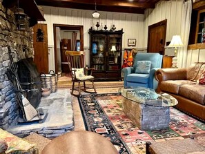 Un salon chaleureux avec une cheminée en pierre, un fauteuil bleu, un canapé en cuir marron et un tapis à motifs.