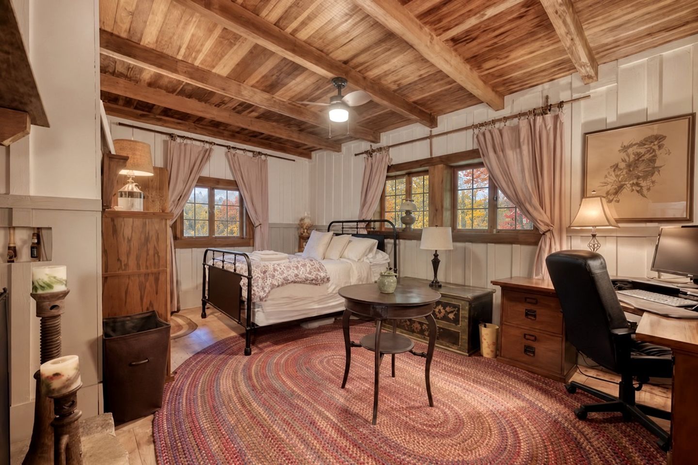 Una habitación con cama, escritorio, silla y una mesa redonda. Hay ventanas con cortinas, un ventilador de techo y un cuadro en la pared.