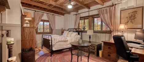 Una habitación con cama, escritorio, silla y una mesa redonda. Hay ventanas con cortinas, un ventilador de techo y un cuadro en la pared.