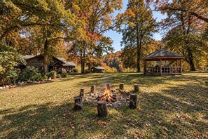 A campsite with a fire pit, wooden cabins, and a gazebo surrounded by trees with autumn foliage.