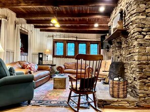 A cozy living room with a stone fireplace, wooden ceiling beams, and a large window with multiple panes. - Newly Restored Cabin in Dahlonega, Georgia, Surrounded by Wineries and Cozy Nature (Dahlonega)