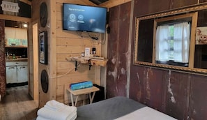 A wooden cabin room with a bed, a television mounted on the wall, a small table, and a window with curtains. - Pet-Friendly Tiny Home Rental in the Chattahoochee National Forest, for Glamping in Georgia (Suches)