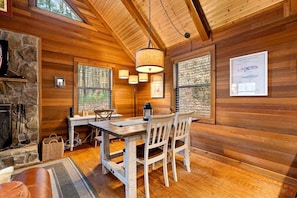 A wooden cabin interior with a dining table, chairs, a hanging light fixture, a stone fireplace, and framed pictures on the walls.