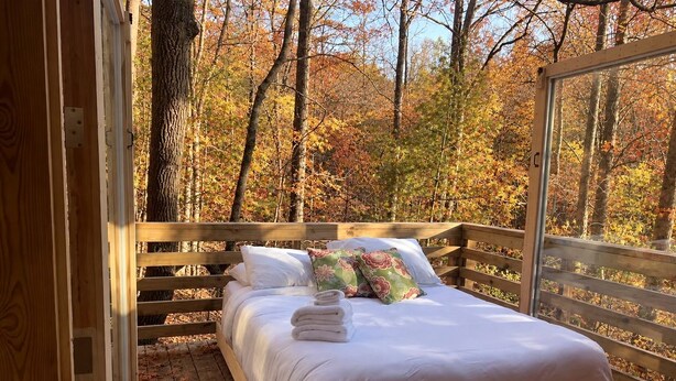 A wooden bed with white bedding and pillows, set on a wooden deck with a view of autumn trees. - Charming Tree House with a Hot Tub in Chattahoochee National Forest, Georgia (Suches)