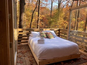 Una cama de madera con ropa blanca y almohadas, ubicada en una terraza de madera con vistas a árboles otoñales.