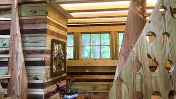 A rustic cabin interior with wooden walls, a framed picture, a table with a vase, and a window with a view of trees.