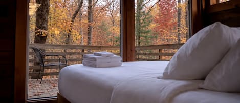 A bed with white linens and pillows, a wooden bench outside the window, and a view of autumn trees.