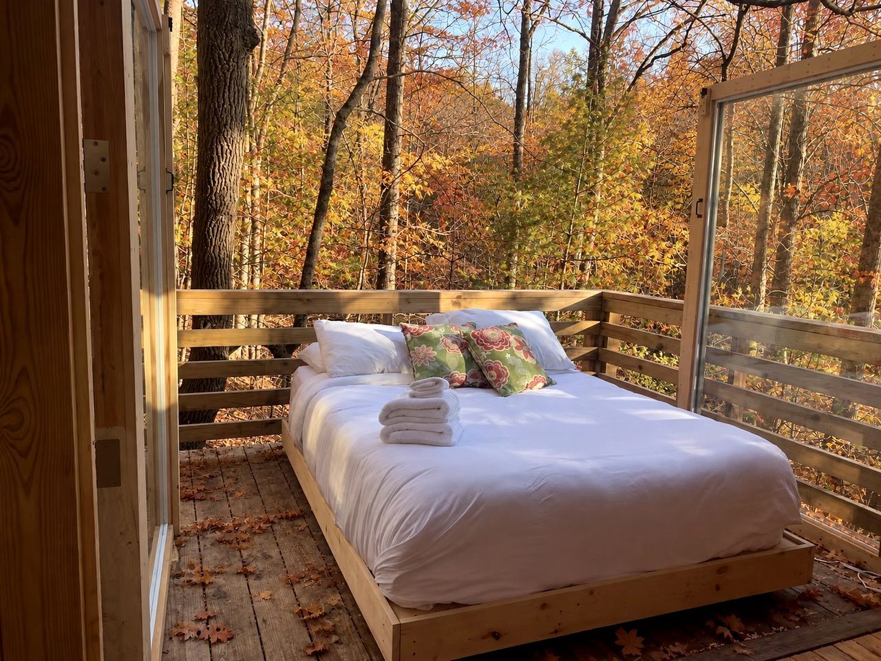 A wooden bed with white bedding and pillows, set on a wooden deck with a view of autumn trees.