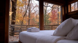 A bed with white linens and pillows, a wooden bench outside the window, and a view of autumn trees. - Charming Tree House with a Hot Tub in Chattahoochee National Forest, Georgia (Suches)