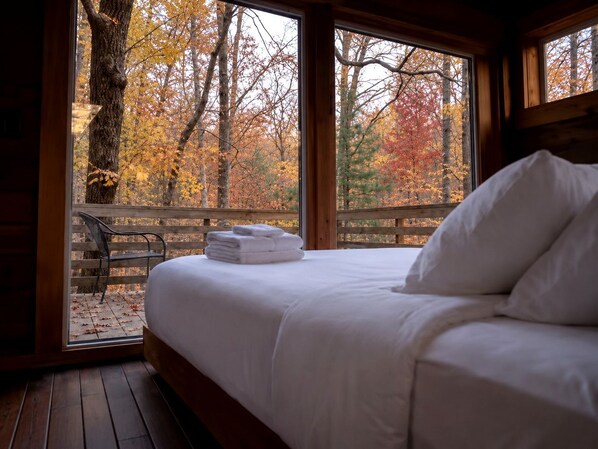 A bed with white linens and pillows, a wooden bench outside the window, and a view of autumn trees.