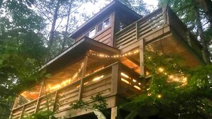 Een houten boomhuis met meerdere verdiepingen, sfeerverlichting en uitzicht op het omliggende bos.