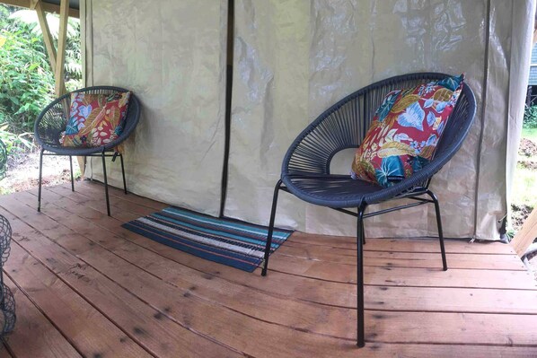 Une véranda couverte avec deux fauteuils en osier, chacun agrémenté d’un coussin coloré, et un tapis à rayures sur une terrasse en bois.