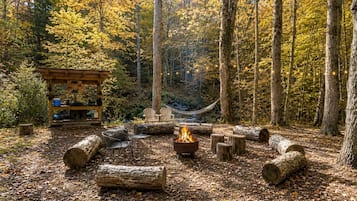 Un camping en el bosque con una fogata, asientos y una pequeña construcción de madera.