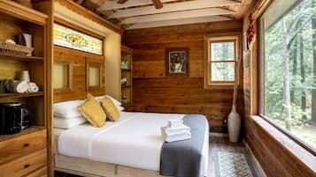 A wooden cabin bedroom with a bed, shelves, a coffee maker, a window with a view of trees, and a ceiling fan.