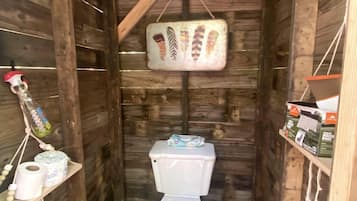A wooden cabin-style bathroom with a toilet, a hanging toilet paper holder, and a shelf with toiletries.
