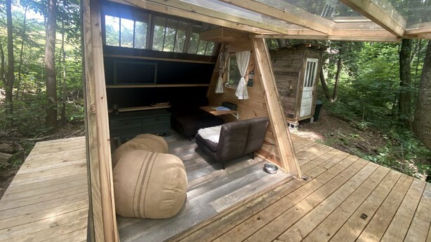 A wooden deck with a sofa and ottoman, a small table, and a window overlooking a forest. - Off-Grid Experience in Cozy Mini A-Frame in Picturesque Tennessee (Grandview)