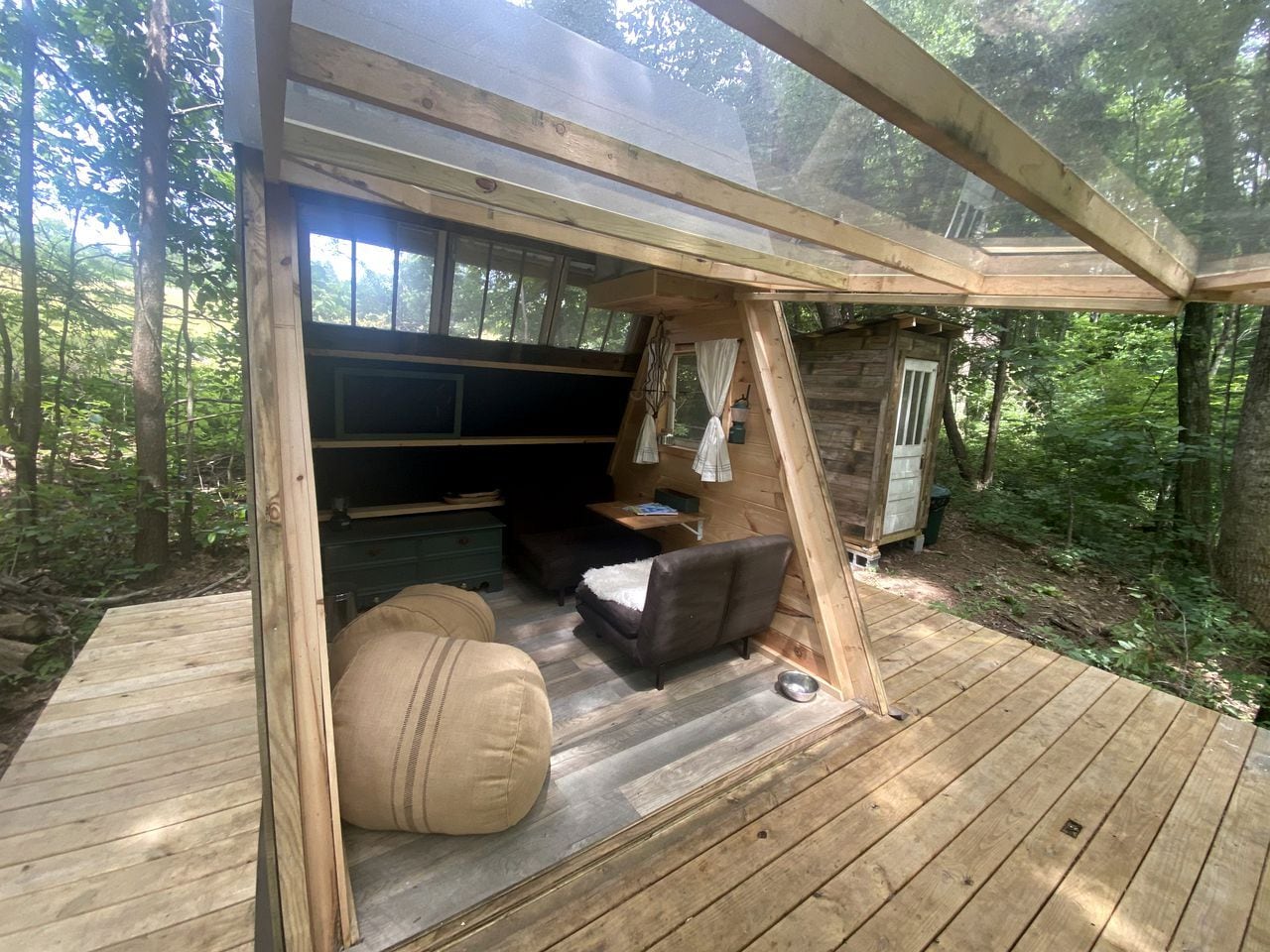 A wooden deck with a sofa and ottoman, a small table, and a window overlooking a forest.