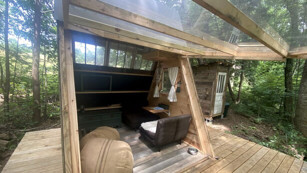 Terraza de madera con sofá, reposapiés, una mesita y ventana con vistas a un bosque.