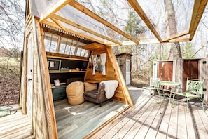 Une cabane en bois avec une chambre à coucher en mezzanine, un coin repas avec des sièges extérieurs et un petit abri de jardin, le tout situé dans un cadre boisé.