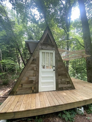 A wooden triangular cabin with a white door, surrounded by a wooden deck and forest.