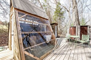 A wooden structure with a large window, a small deck with a table and chairs, and a shed in the background. - Off-Grid Experience in Cozy Mini A-Frame in Picturesque Tennessee (Grandview)