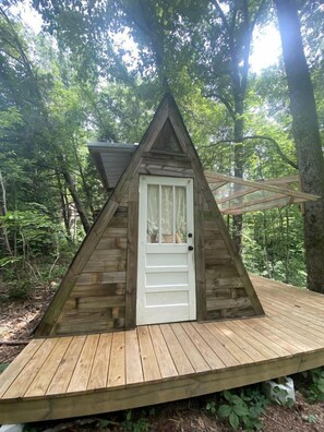A wooden triangular cabin with a white door, surrounded by a wooden deck and forest. - Off-Grid Experience in Cozy Mini A-Frame in Picturesque Tennessee (Grandview)