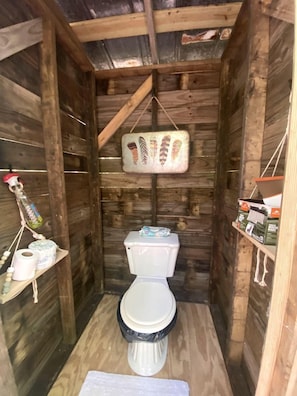 Une salle de bain de style rustique, avec des murs en bois, comprenant des toilettes, un porte-rouleau de papier toilette suspendu et une étagère avec des articles de toilette.
