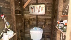 A wooden cabin-style bathroom with a toilet, a hanging toilet paper holder, and a shelf with toiletries. - Off-Grid Experience in Cozy Mini A-Frame in Picturesque Tennessee (Grandview)