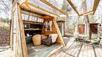 Uma cabana de madeira com quarto no mezanino, área de refeições com assentos externos e um pequeno galpão em um ambiente arborizado.