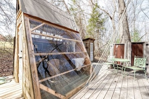 Une structure en bois dotée d’une grande fenêtre, d’une petite terrasse avec une table et des chaises, et d’un abri de jardin en arrière-plan.