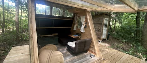 Eine Holzterrasse mit einer Couch, einem Hocker, einem kleinen Tisch und einem Fenster mit Blick auf einen Wald.