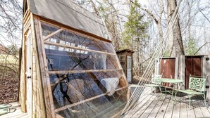 A wooden structure with a large window, a small deck with a table and chairs, and a shed in the background. - Off-Grid Experience in Cozy Mini A-Frame in Picturesque Tennessee (Grandview)