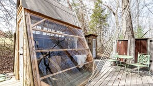 A wooden structure with a large window, a small deck with a table and chairs, and a shed in the background. - Off-Grid Experience in Cozy Mini A-Frame in Picturesque Tennessee (Grandview)