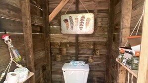 A wooden cabin-style bathroom with a toilet, a hanging toilet paper holder, and a shelf with toiletries. - Off-Grid Experience in Cozy Mini A-Frame in Picturesque Tennessee (Grandview)