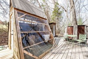 A wooden structure with a large window, a small deck with a table and chairs, and a shed in the background. - Off-Grid Experience in Cozy Mini A-Frame in Picturesque Tennessee (Grandview)