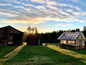 A barn, a small shed, and a greenhouse in a grassy field with a sunset in the background.